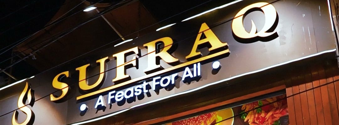 The lit Sufraq shopfront sign at night, reading Sufraq, A Feast For All